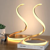Modern gold spiral table lamp on a wooden surface with a plant and decorative deer figurine.