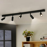 Black ceiling track light fixture with four spotlights in a room with a window and bookshelf.