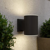 Black outdoor wall light fixture on a concrete wall with plants around.