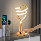 Gold spiral table lamp on a desk with a hand plugging in a device, smartphone, and smartwatch in the background.