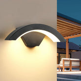 Modern outdoor wall light fixture on a house exterior with patio furniture in the background.