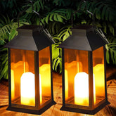 Two black lanterns with glowing candles on a wooden surface with green foliage in the background