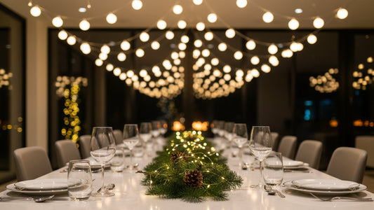 Cozy Christmas dinner table under warm festoon lights with candles, pine branches, ornaments, and glowing tree in background."