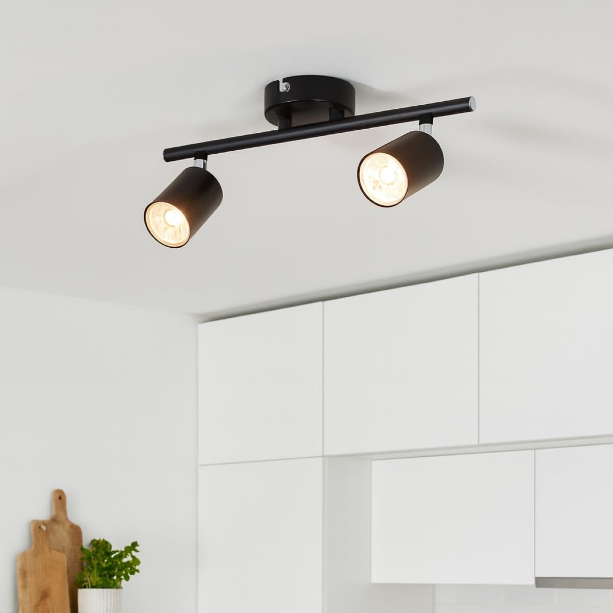 Black ceiling light fixture with two spotlights on a white ceiling above kitchen cabinets.