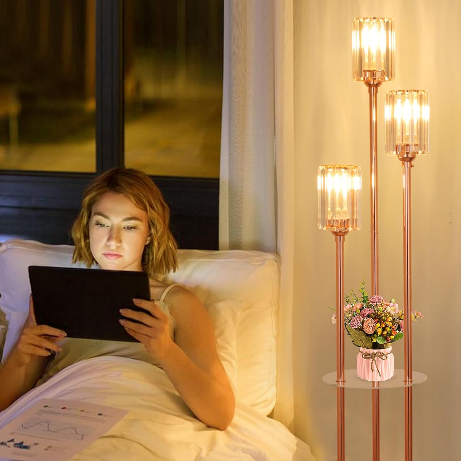 Woman lying in bed using a tablet with a cozy room setting featuring floor lamps and a vase of flowers.