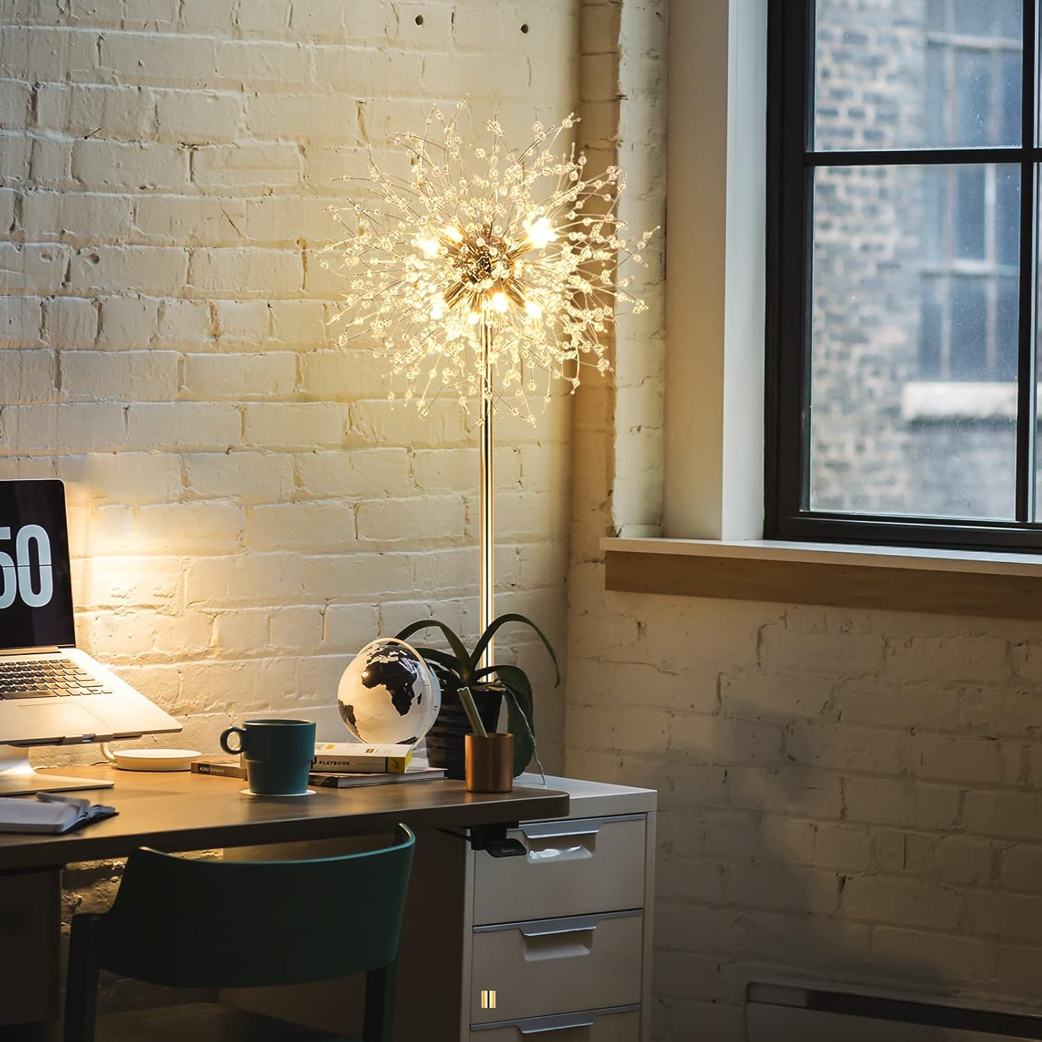 Modern office desk with a laptop, mug, and decorative lamp against a brick wall.