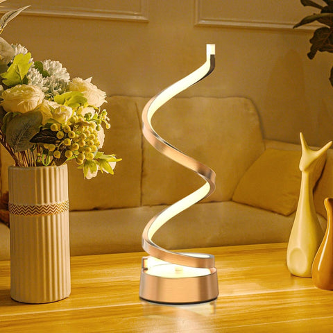 Spiral table lamp on a wooden surface with flowers and a vase in the background