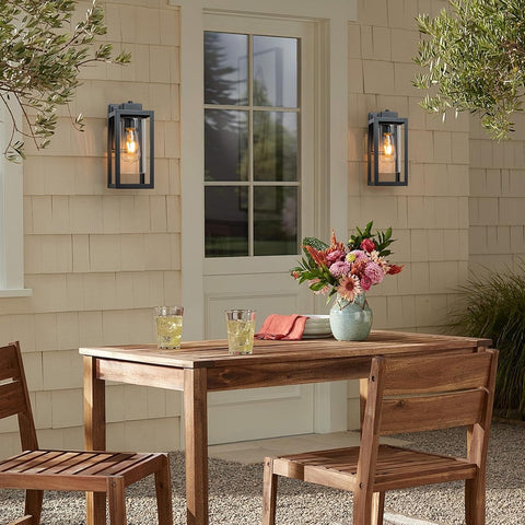 Wooden outdoor table with chairs, glasses, and a vase of flowers against a beige wall.