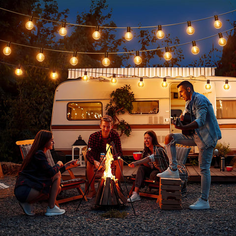 Group of people around a campfire with a camper van in the background under string lights.
