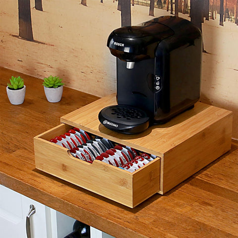 Black coffee machine on a wooden stand with a drawer containing coffee pods, set against a wooden wall.