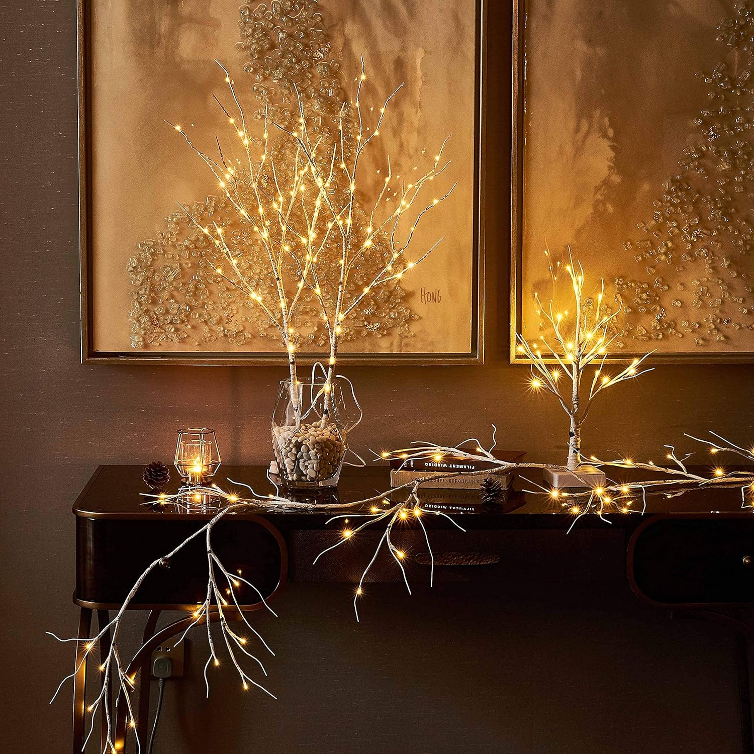 Decorative setup with LED lights on branches, candles, and books against a brown wall with framed artwork.