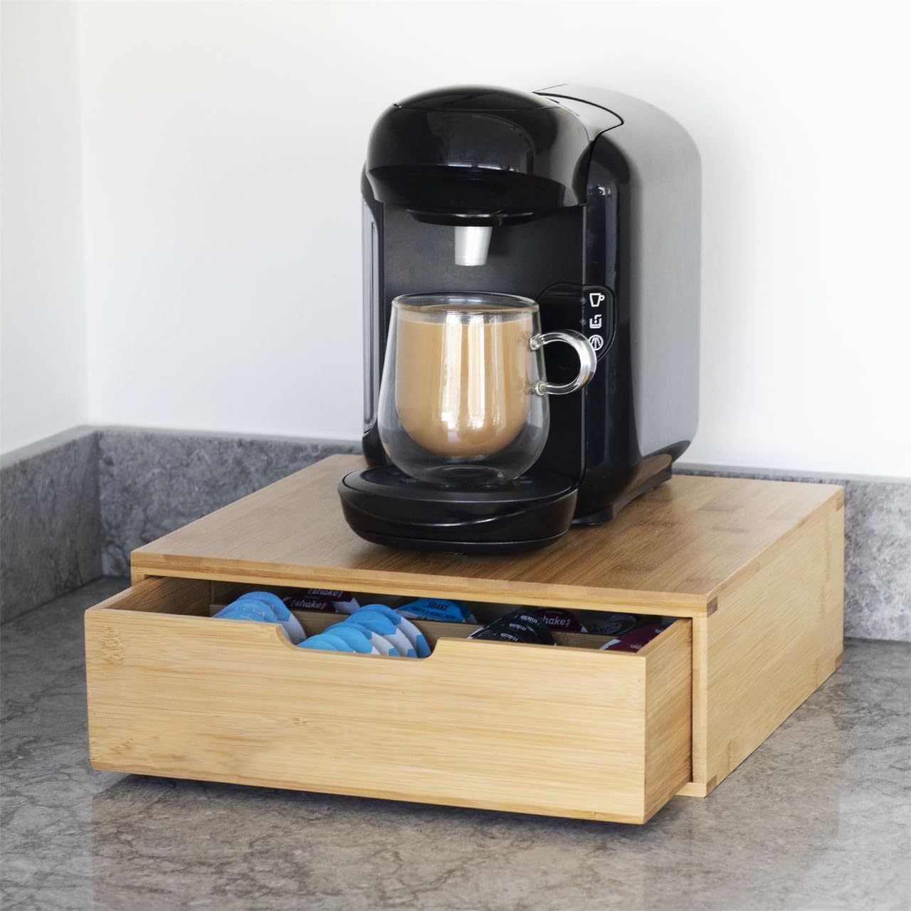 Black coffee machine with a glass mug on a wooden stand against a white wall.