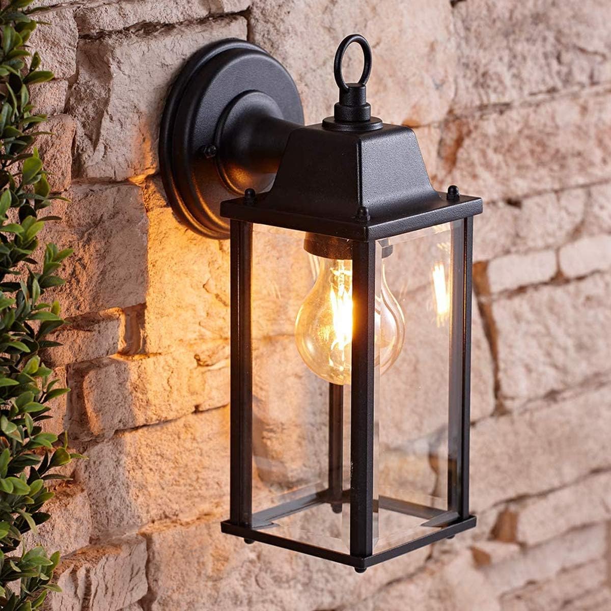 Black outdoor wall lantern with a lit bulb attached to a stone wall.