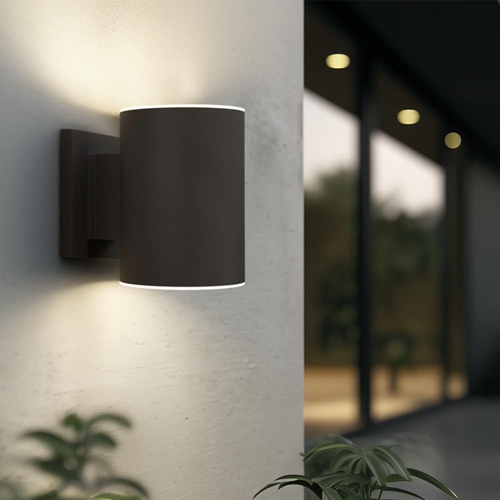 Black outdoor wall light fixture on a light gray wall with plants and blurred background.