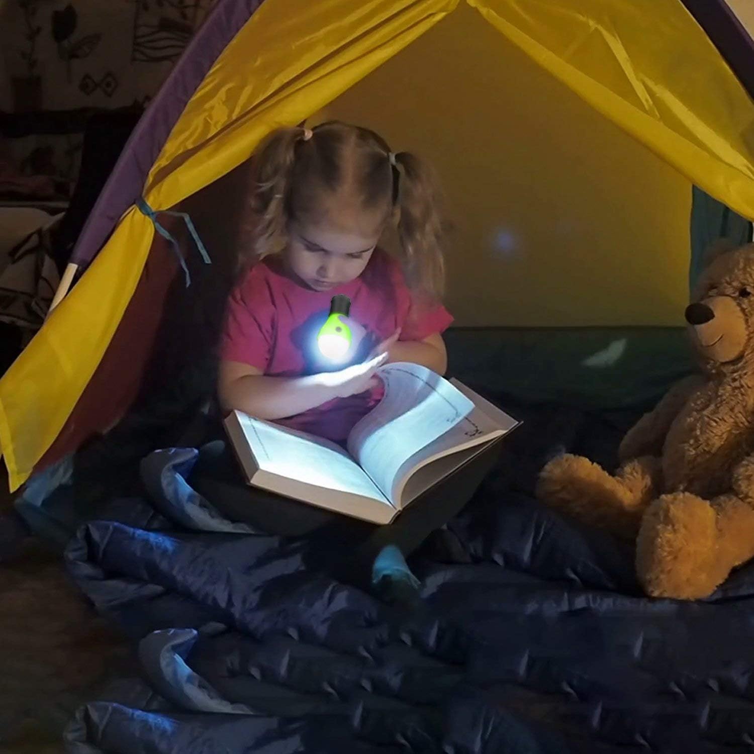 Child reading a book with a flashlight inside a tent, accompanied by a teddy bear.