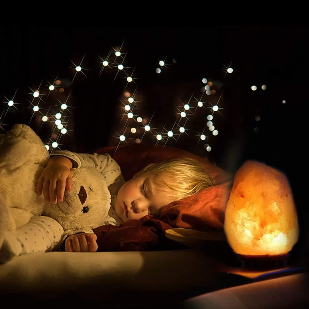 Child sleeping with a teddy bear under string lights, next to a glowing lamp.