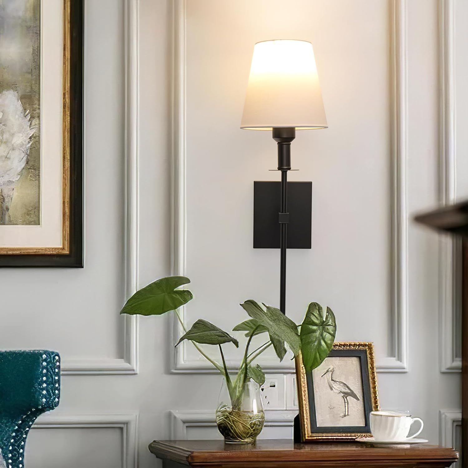 Decorative wall lamp with a plant and framed picture on a table against a paneled wall.