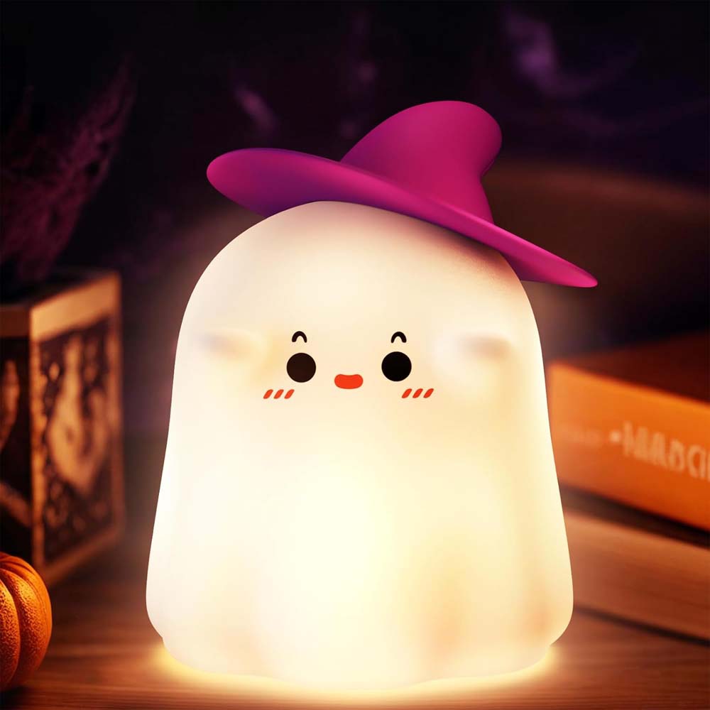 Ghost-shaped night light with a pink witch hat on a wooden surface with Halloween decorations.