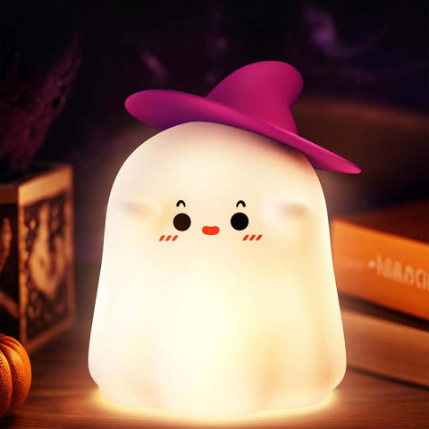 Ghost-shaped night light with a pink witch hat on a wooden surface with Halloween decorations.