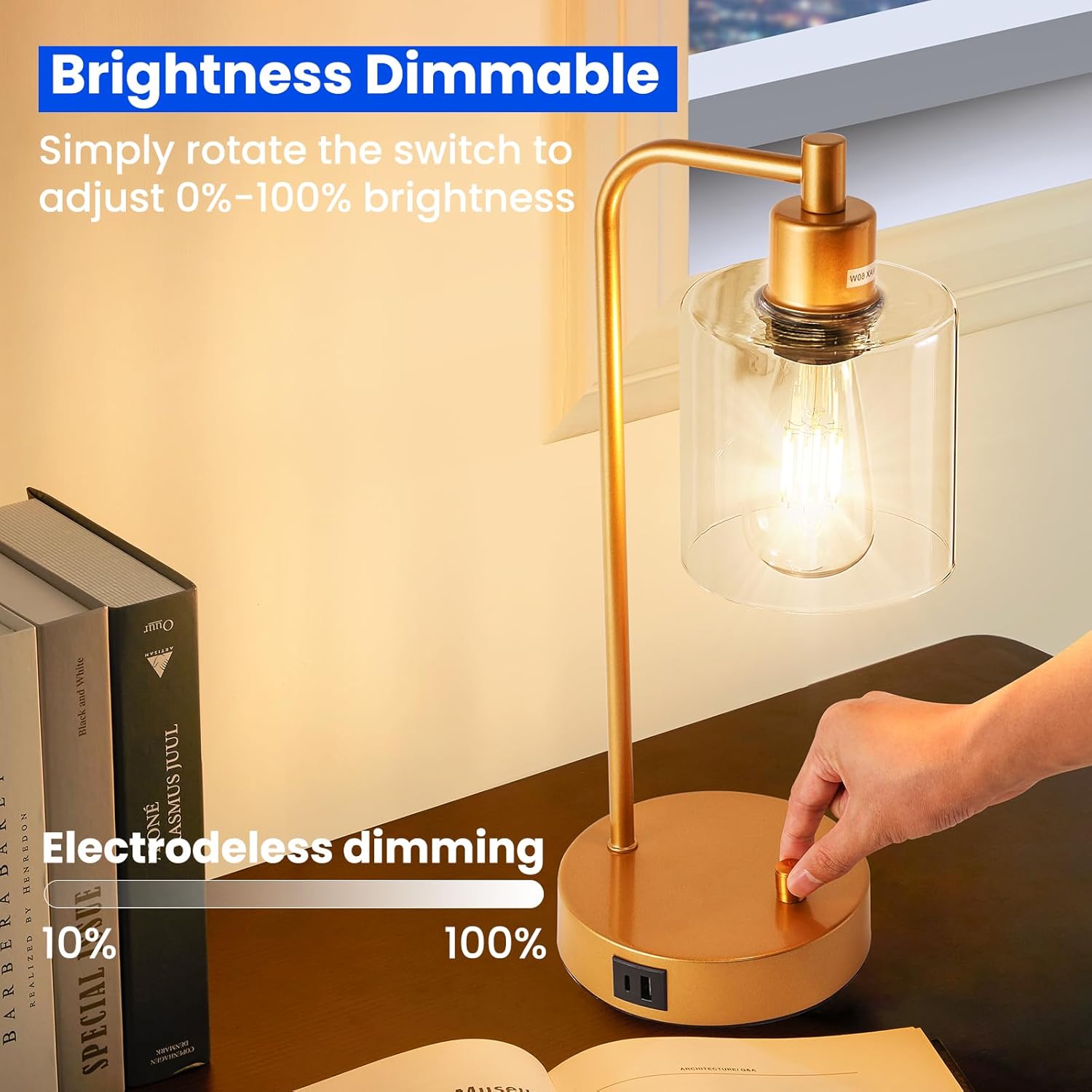 Gold desk lamp with glass shade on a desk next to books, with a hand adjusting the brightness switch.