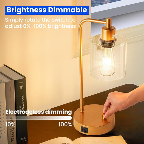 Gold desk lamp with glass shade on a desk next to books, with a hand adjusting the brightness switch.