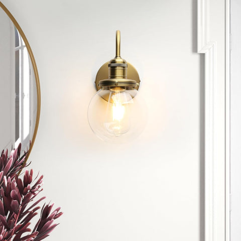 Gold wall-mounted light fixture on a white wall with a plant in the corner.