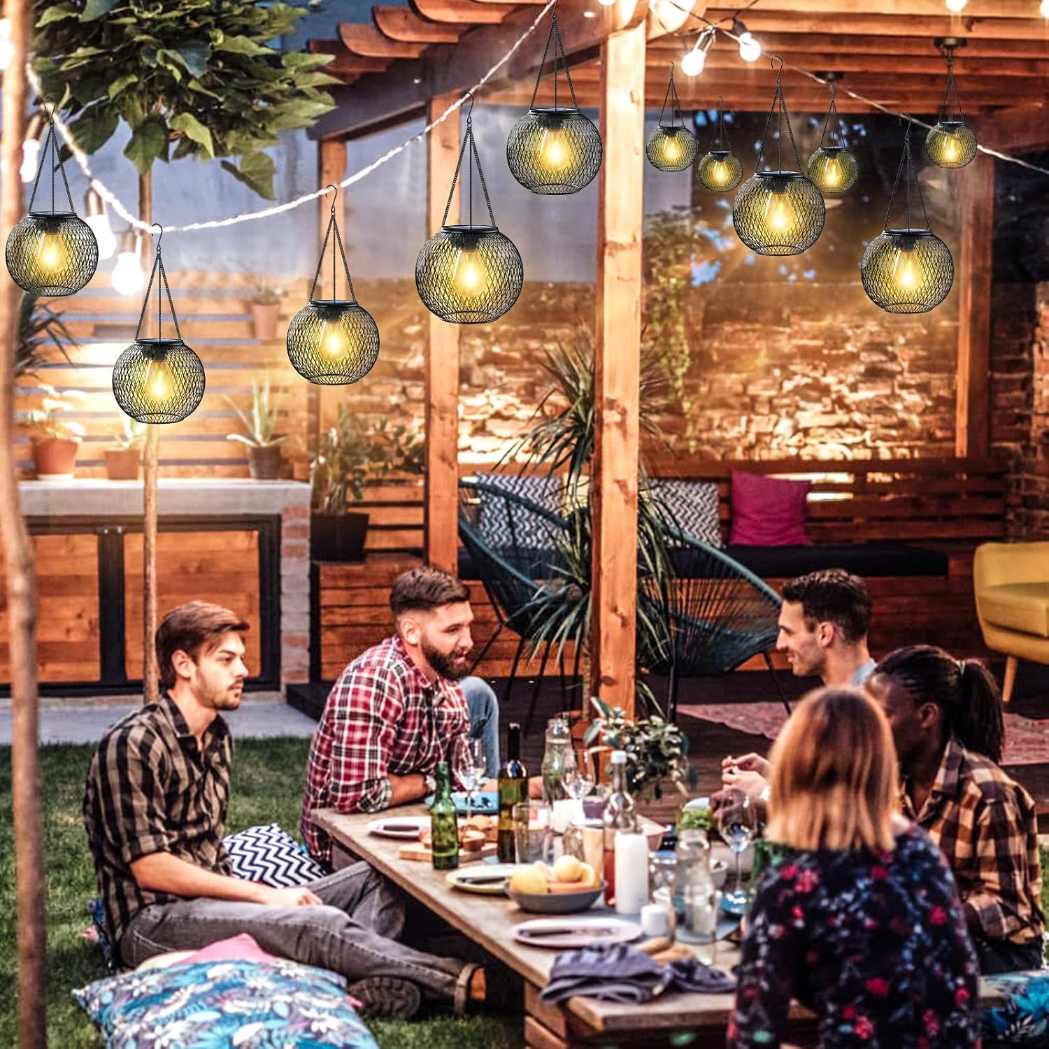 Group of people sitting around a table in an outdoor setting with string lights and plants.