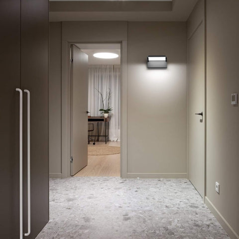 Modern hallway with a light fixture on the wall, open door leading to a room with a desk and plant.