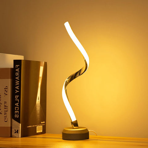 Modern table lamp with curved design on a wooden surface next to books, warm lighting.