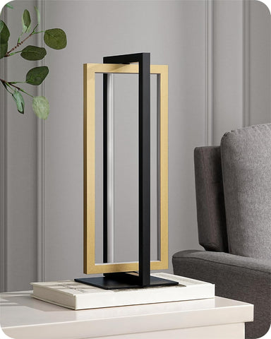 Modern table lamp with black and gold design on a white surface, gray wall, and gray sofa in the background.