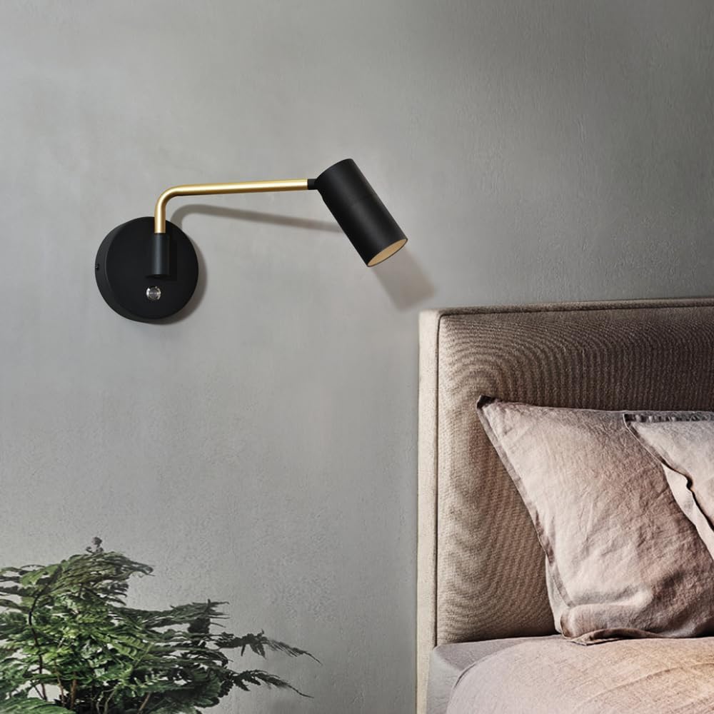 Modern wall lamp with black and gold design on a gray wall next to a bed with pillows.
