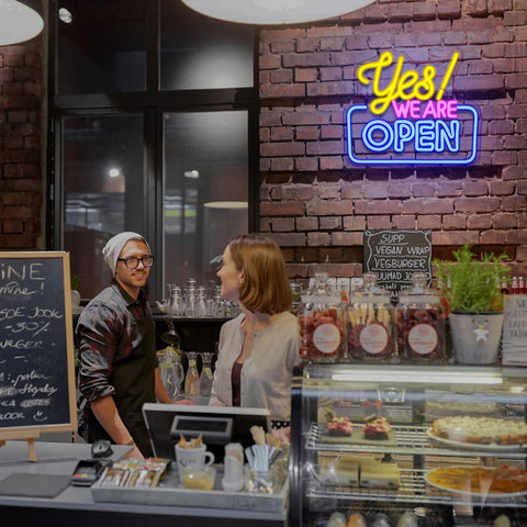 Neon sign 'Yes! We Are Open' above a counter with a customer and employee in a restaurant setting.