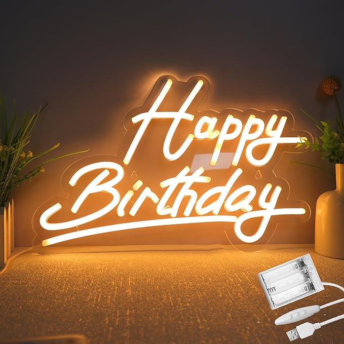Neon sign with 'Happy Birthday' text on a wooden surface with plants and power adapter.