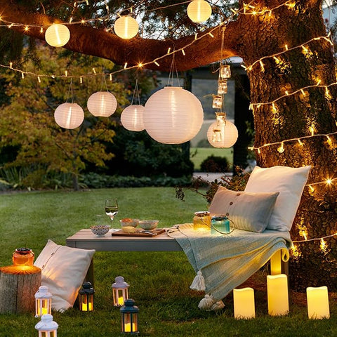 Outdoor setting with string lights, lanterns, and a table with drinks under a tree.