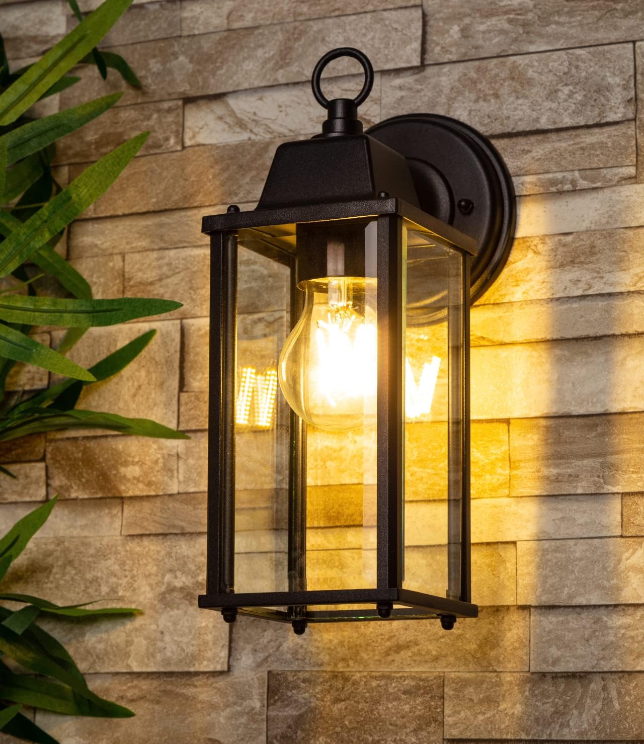 Outdoor wall light fixture on a stone wall with a plant to the left.