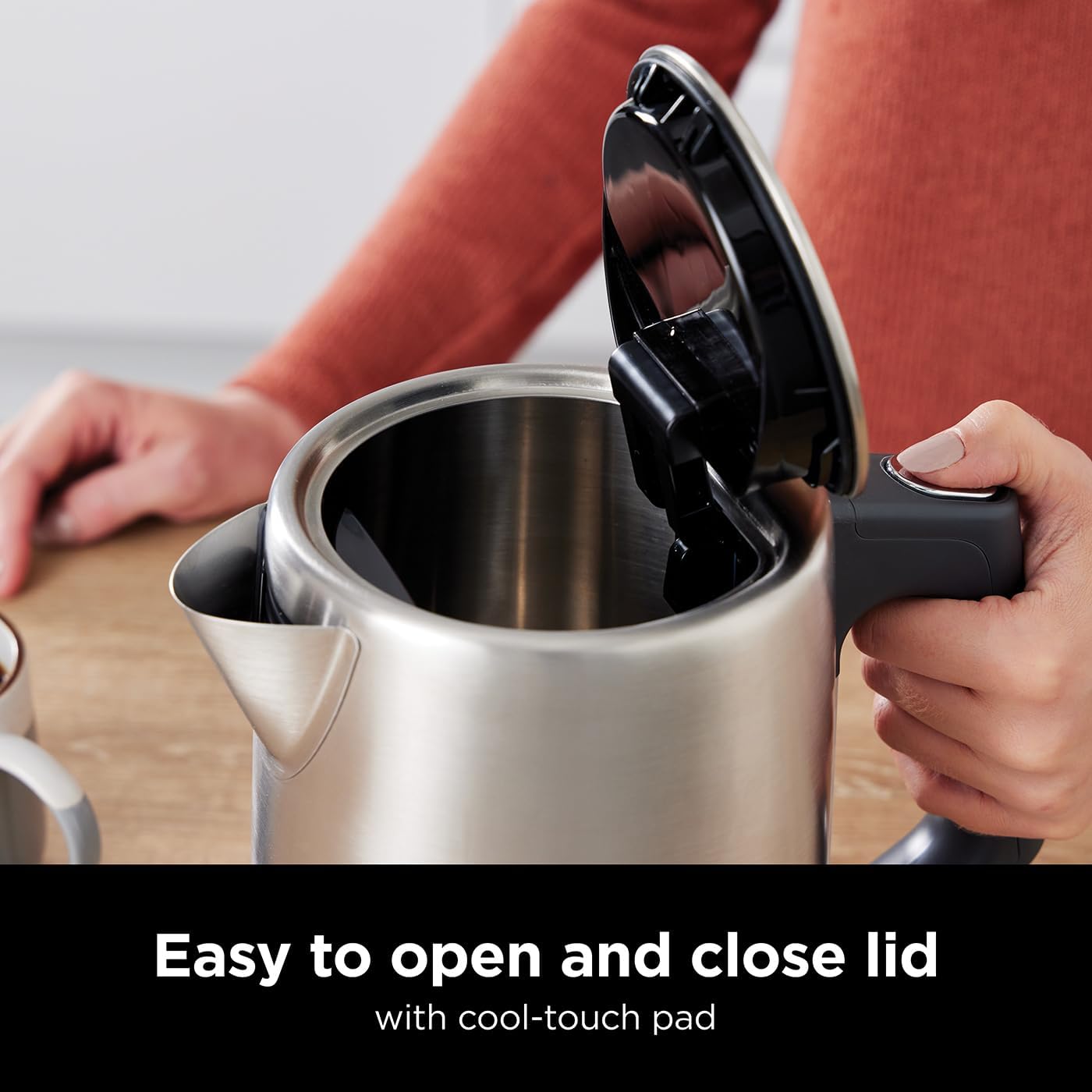 Person opening a silver electric kettle with an easy-to-open lid on a wooden surface.