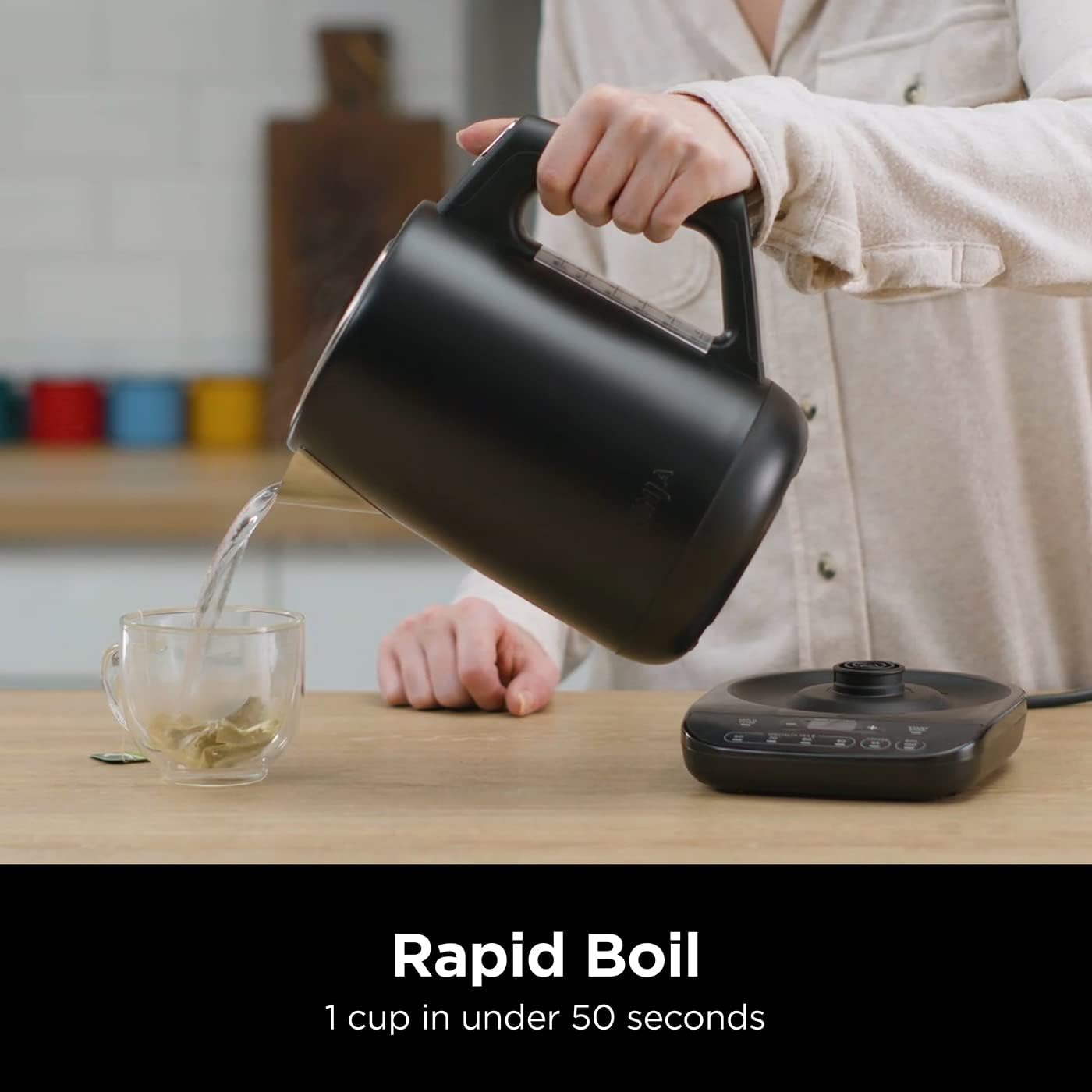 Person pouring tea from a black electric kettle into a glass on a wooden table.