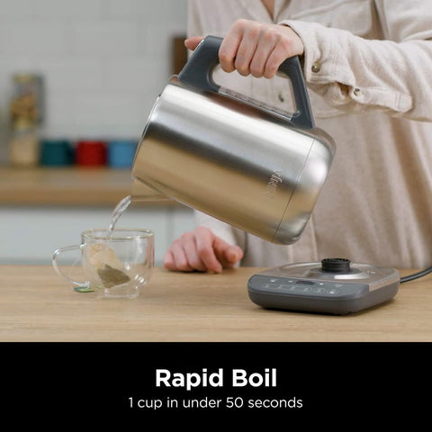 Person pouring tea from a stainless steel kettle into a glass mug on a wooden table.