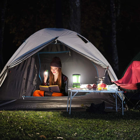 Person reading a book inside a tent with a lantern, surrounded by camping gear.