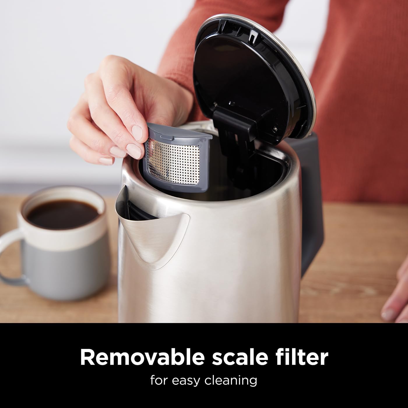 Person removing a removable scale filter from a coffee maker with a cup of coffee on a wooden table.