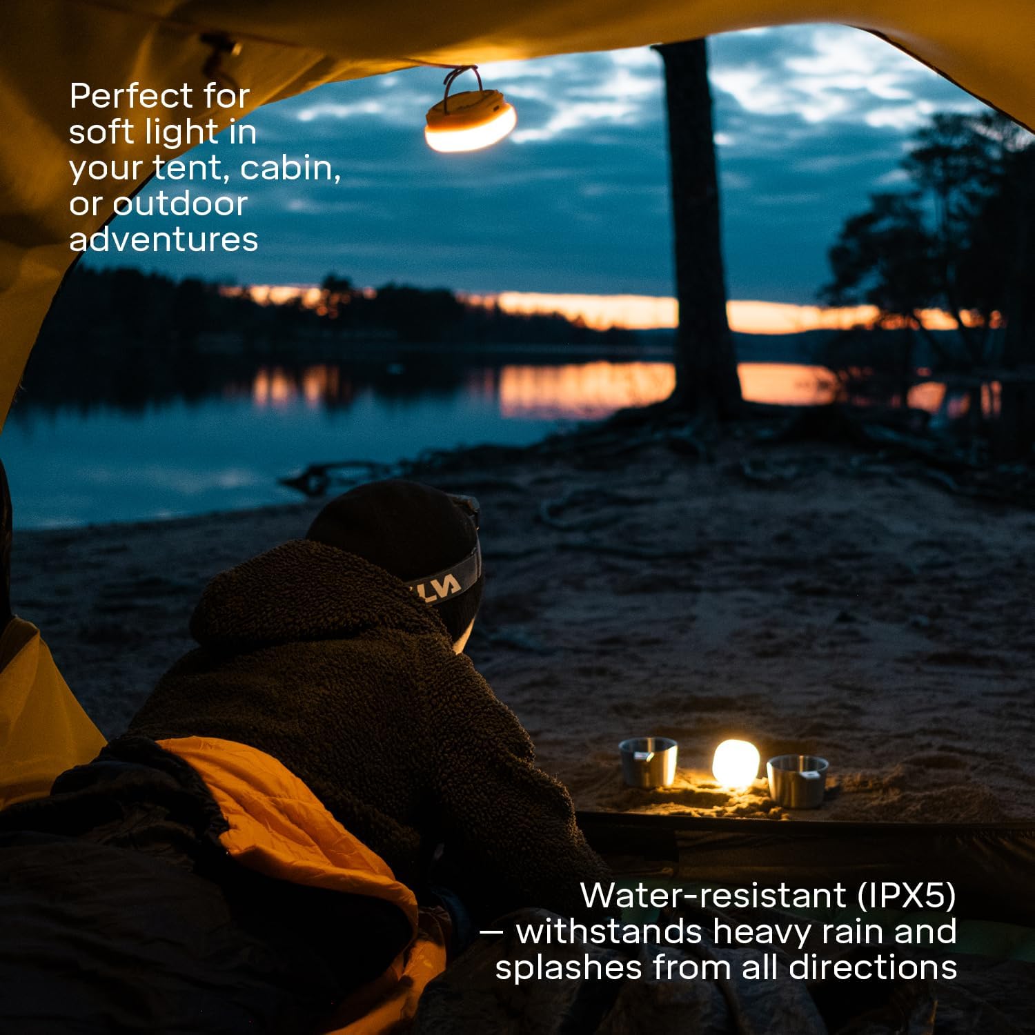 Person sitting under a tent with a lantern, overlooking a lake at dusk.