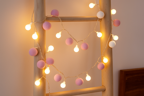 String lights with pink and white bulbs draped over a wooden ladder against a neutral wall.