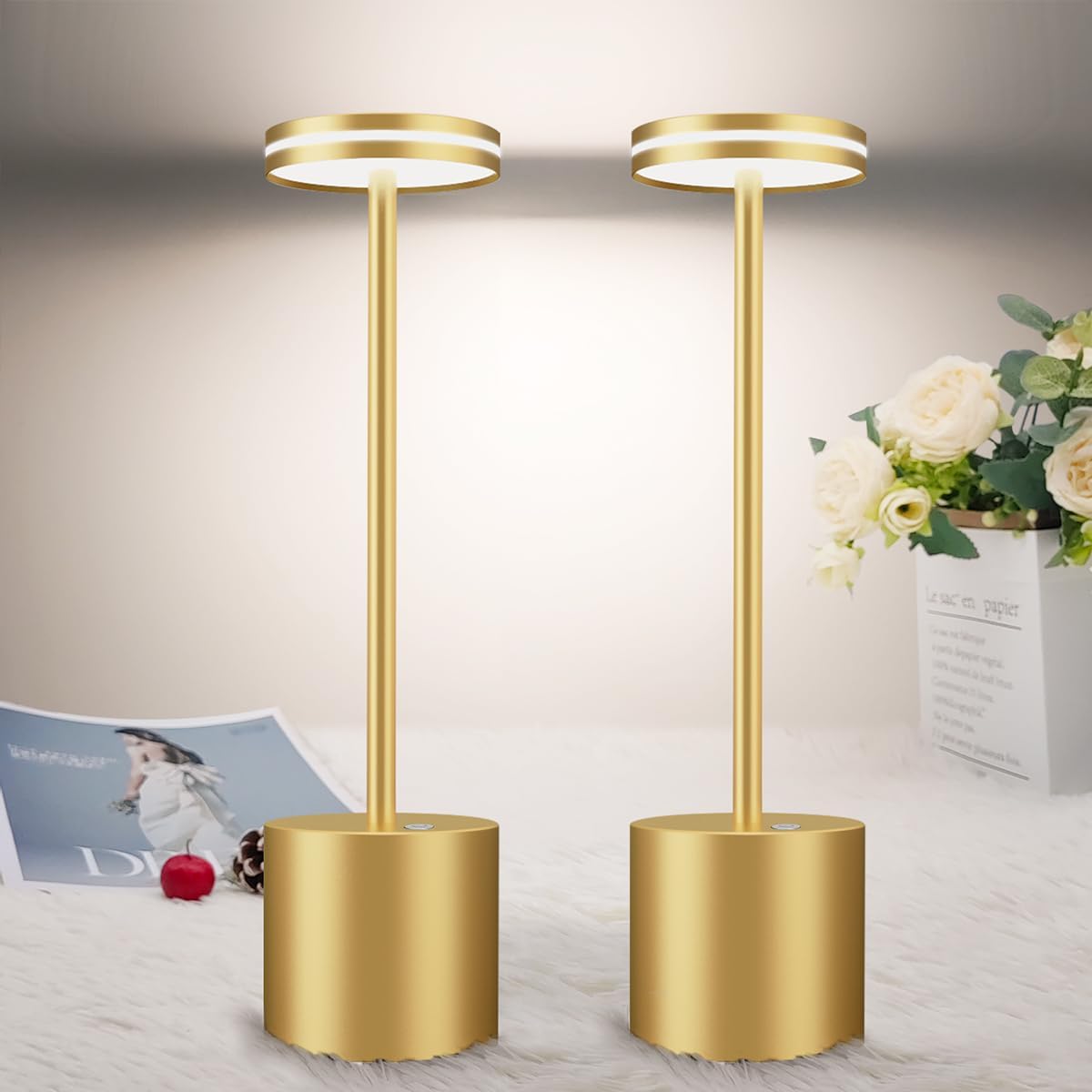 Two gold floor lamps on a light-colored surface with a magazine and flowers in the background.