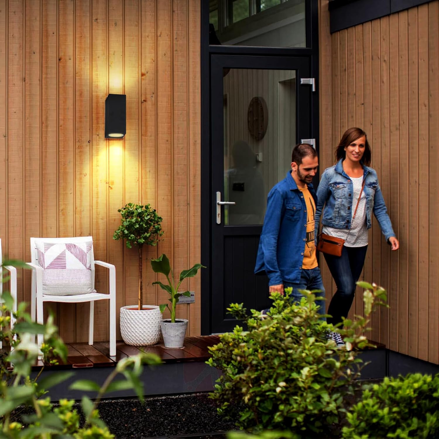 Two people walking towards a modern house entrance with plants and a chair on a wooden deck.