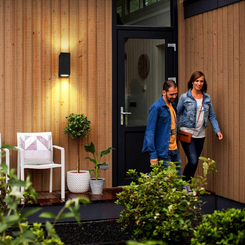 Two people walking towards a modern house entrance with plants and a chair on a wooden deck.