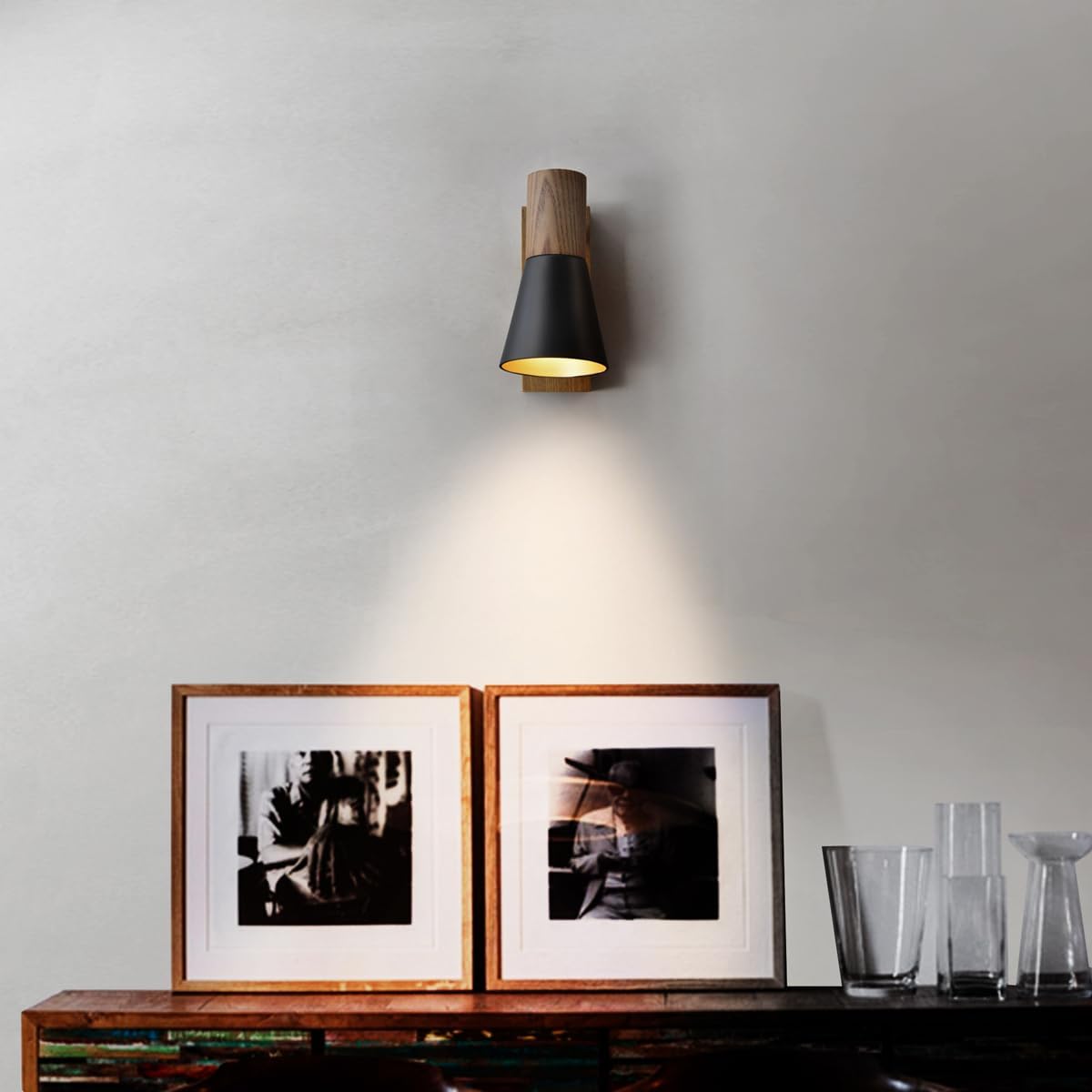 Wall-mounted light fixture casting a warm glow on framed pictures and glassware on a wooden surface.