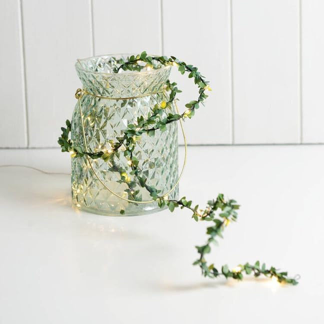 Decorative glass vase with greenery and fairy lights on a white surface