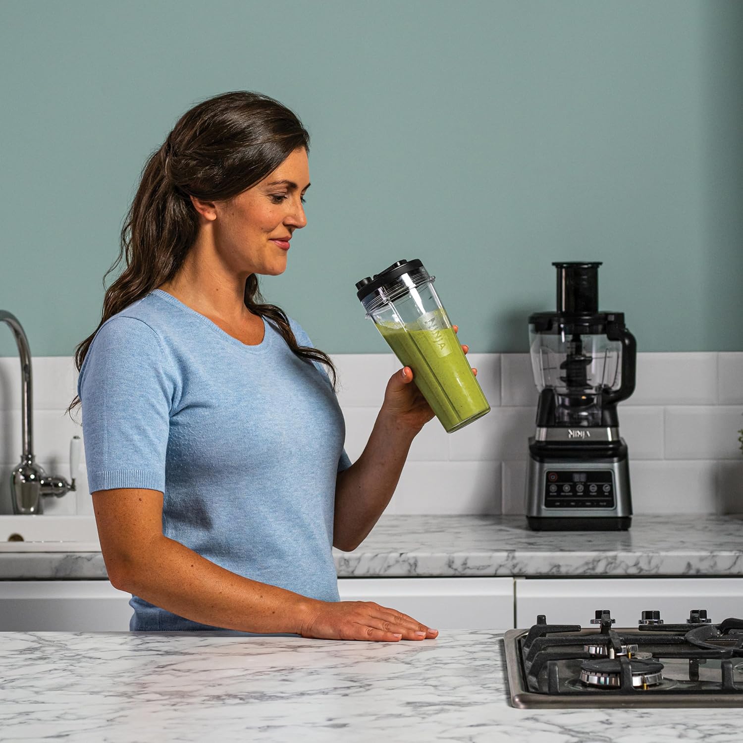Woman holding a green smoothie in a kitchen with a blender and stove.