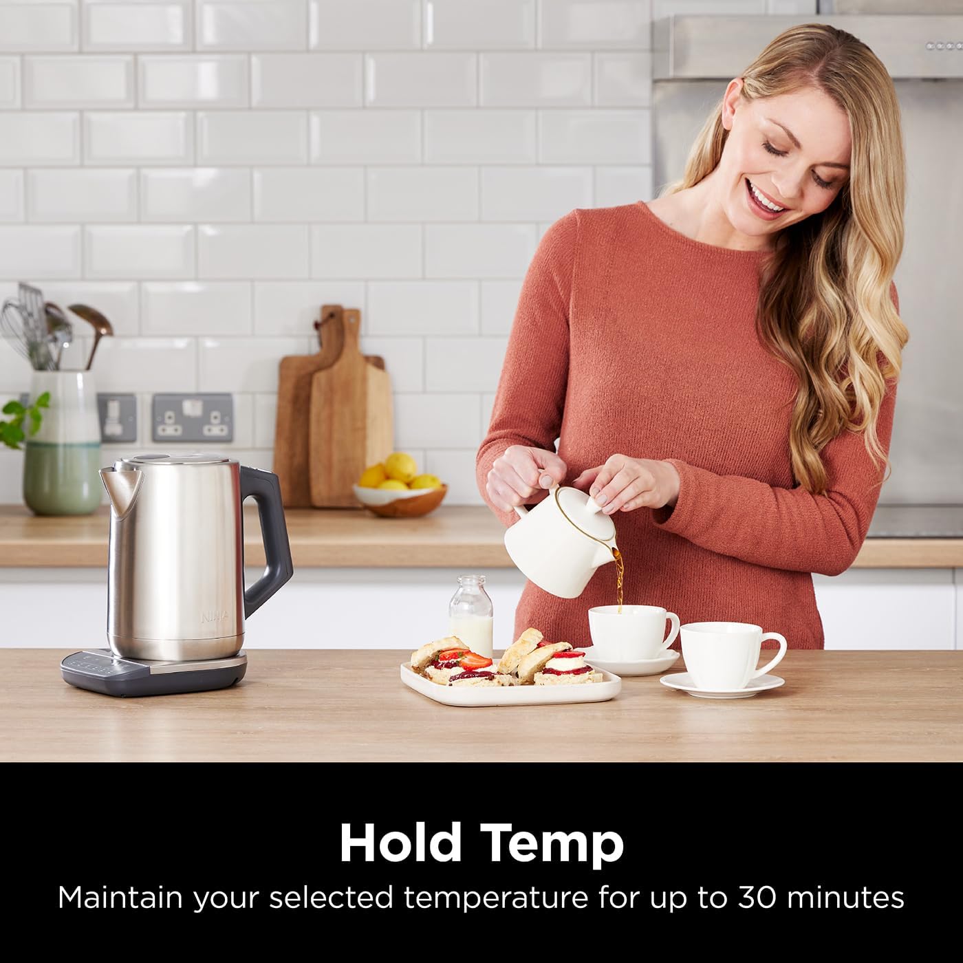 Woman pouring tea into a cup in a kitchen with a kettle and food on the counter.
