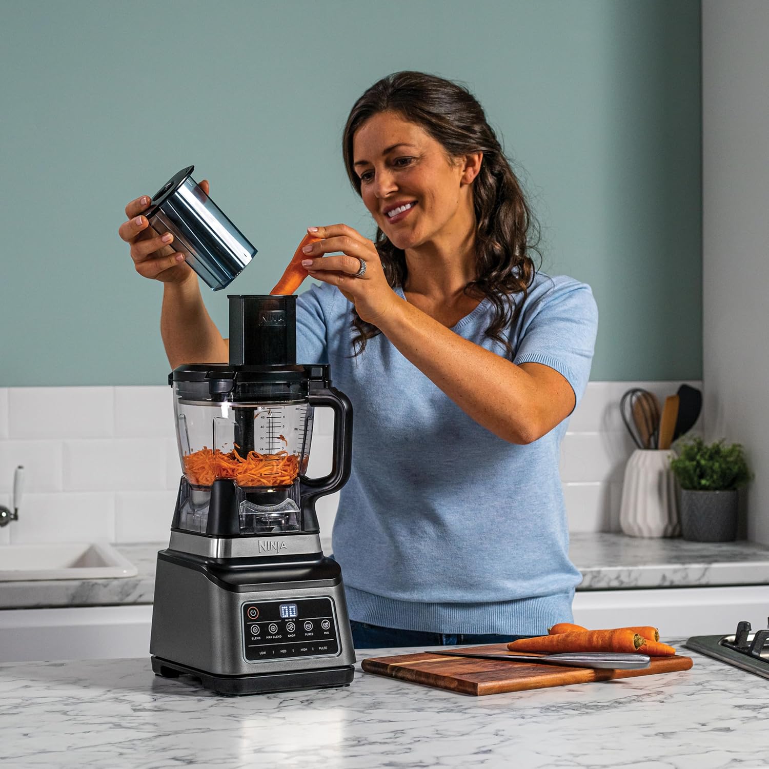 Woman using a blender in a kitchen with carrots on a cutting board.